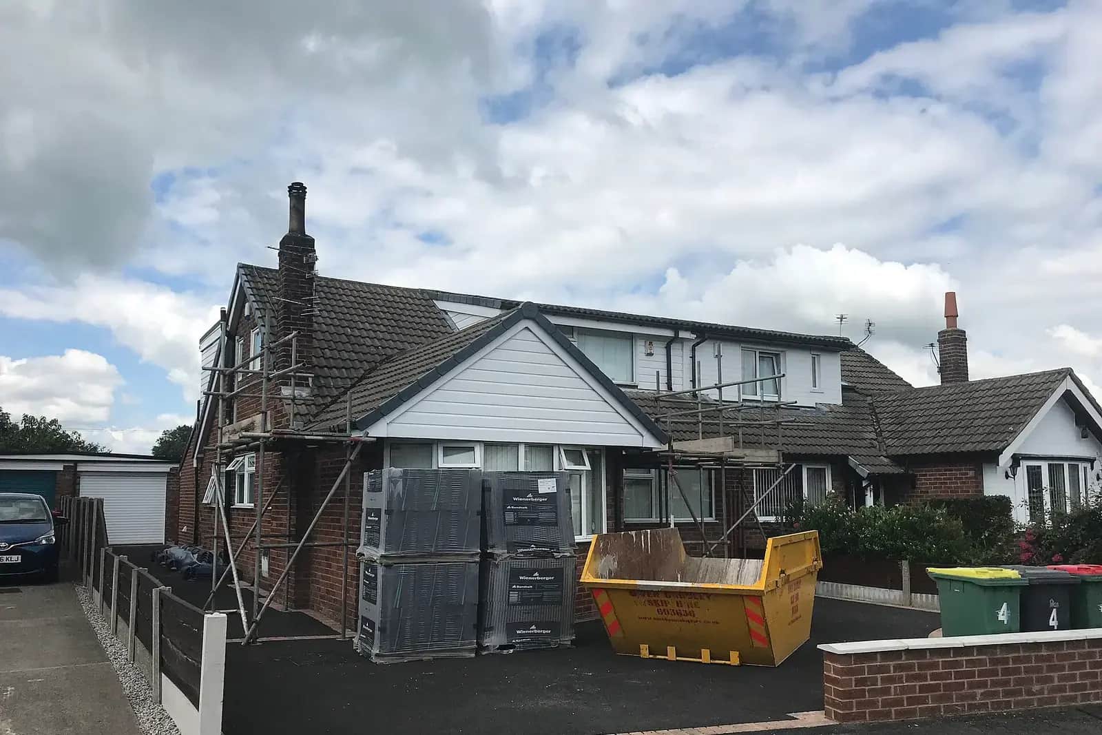 Semi-detached bungalow with a front dormer under construction, scaffolding, stacked roof tiles and a yellow skip on the drive