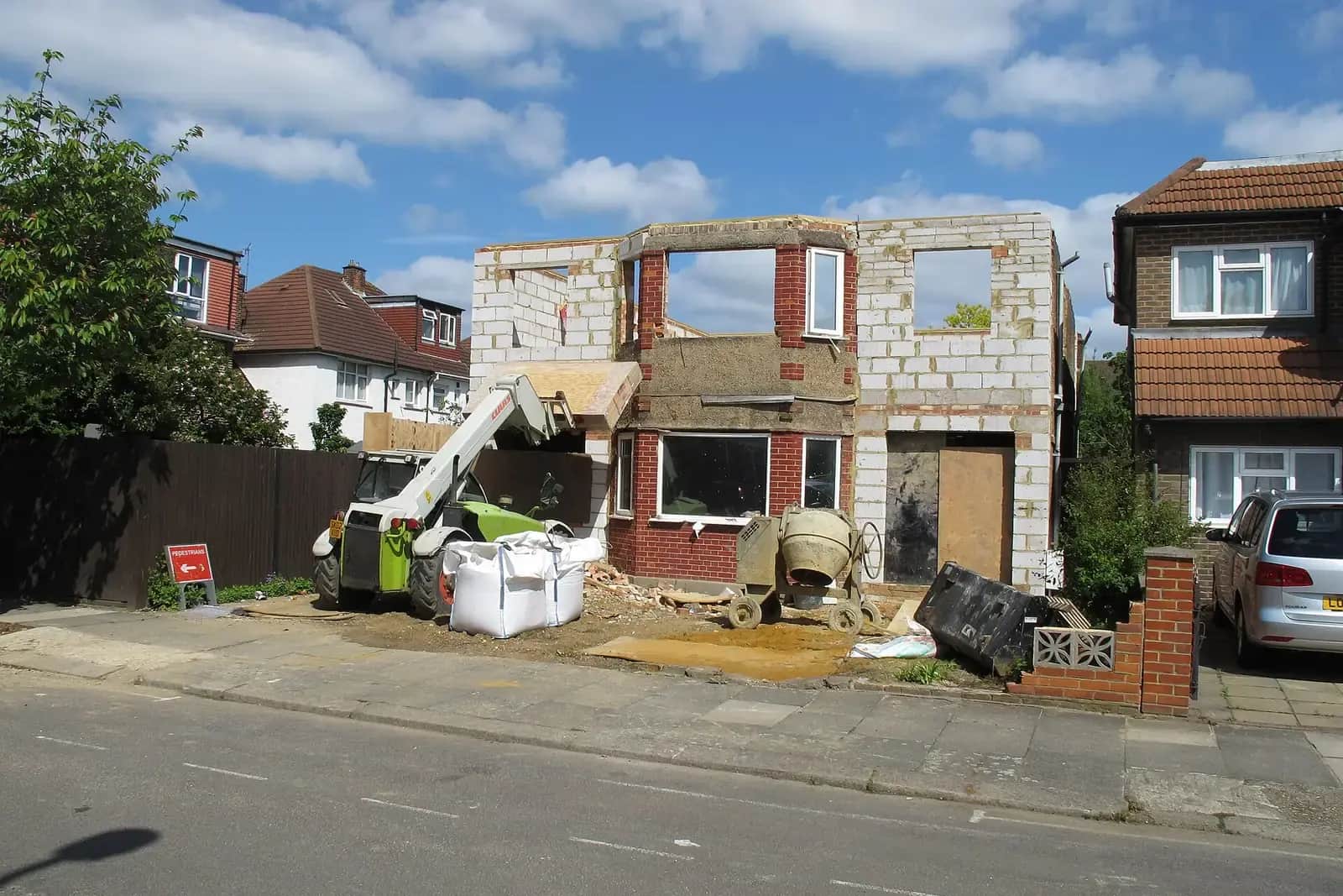 Two-storey extension shell in white blockwork with open window holes beside a semi-detached house, a cement mixer and telehandler in front