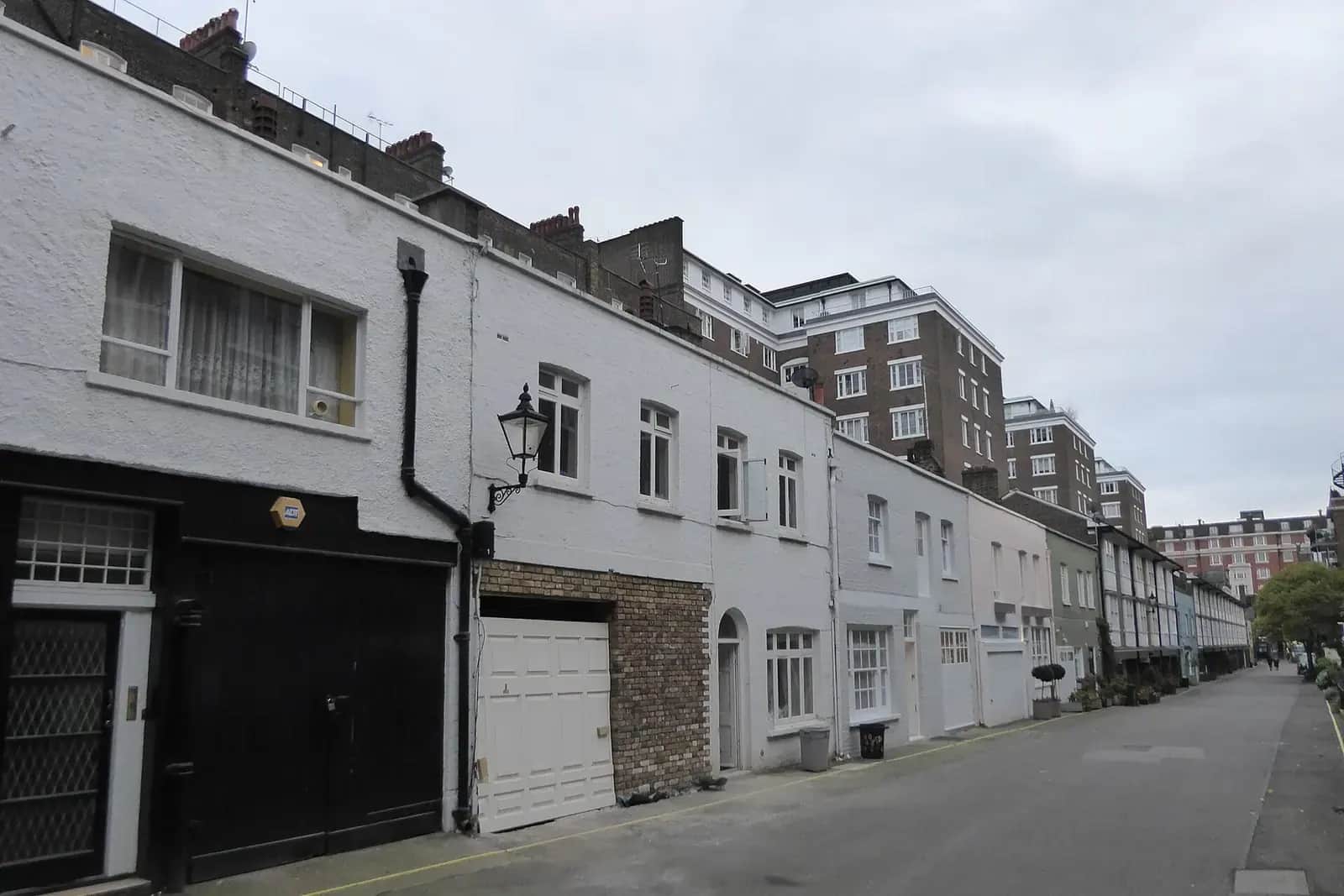 London mews of white-painted converted coach houses with black and white garage doors and a gas-style lamp, mansion flats beyond