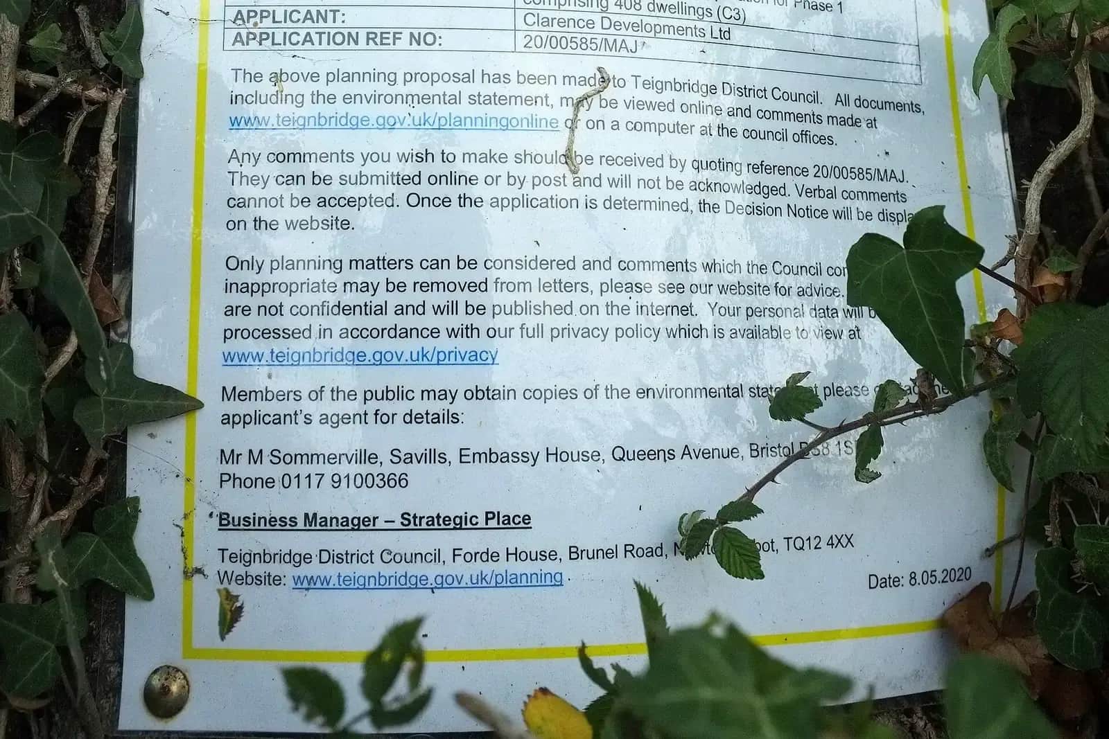 Laminated planning application notice with the council's contact details, partly hidden by ivy on a fence