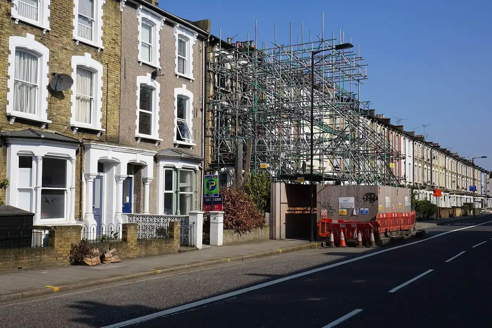 Victorian terrace with a tall freestanding scaffold tower and hoarding on a main road, white-framed sash windows on the houses beside it