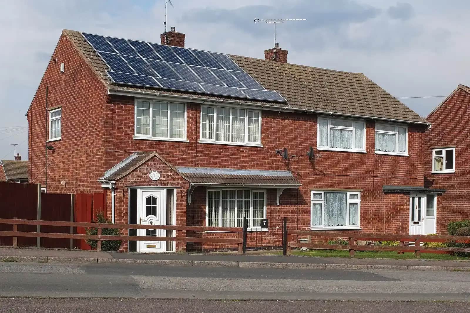 Red-brick semi-detached house with solar panels on its roof, a wooden fence in front and a white front door