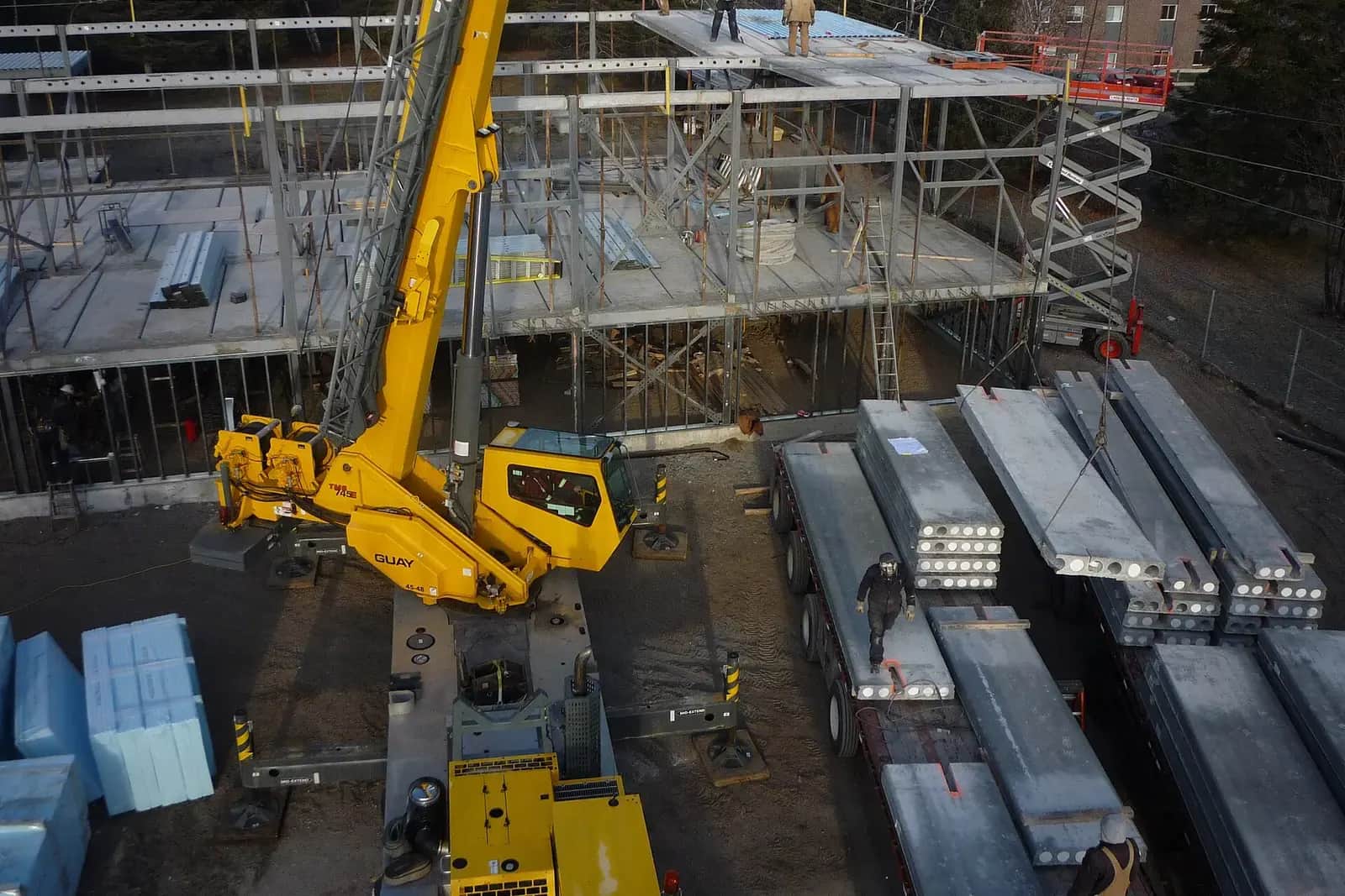 Yellow crane lifting precast concrete floor slabs from a lorry on a building site, a steel frame behind