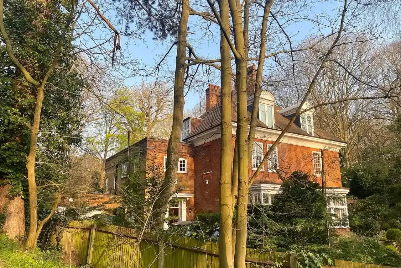 Large red-brick house with dormers seen between tall bare trees on a sunny spring day, a wooden fence in front