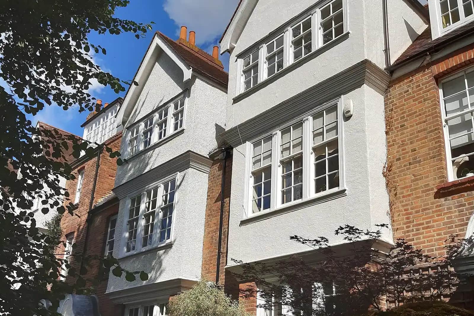 Two tall white roughcast houses with gabled bays and white sash windows between red brick walls, with a Japanese maple in front