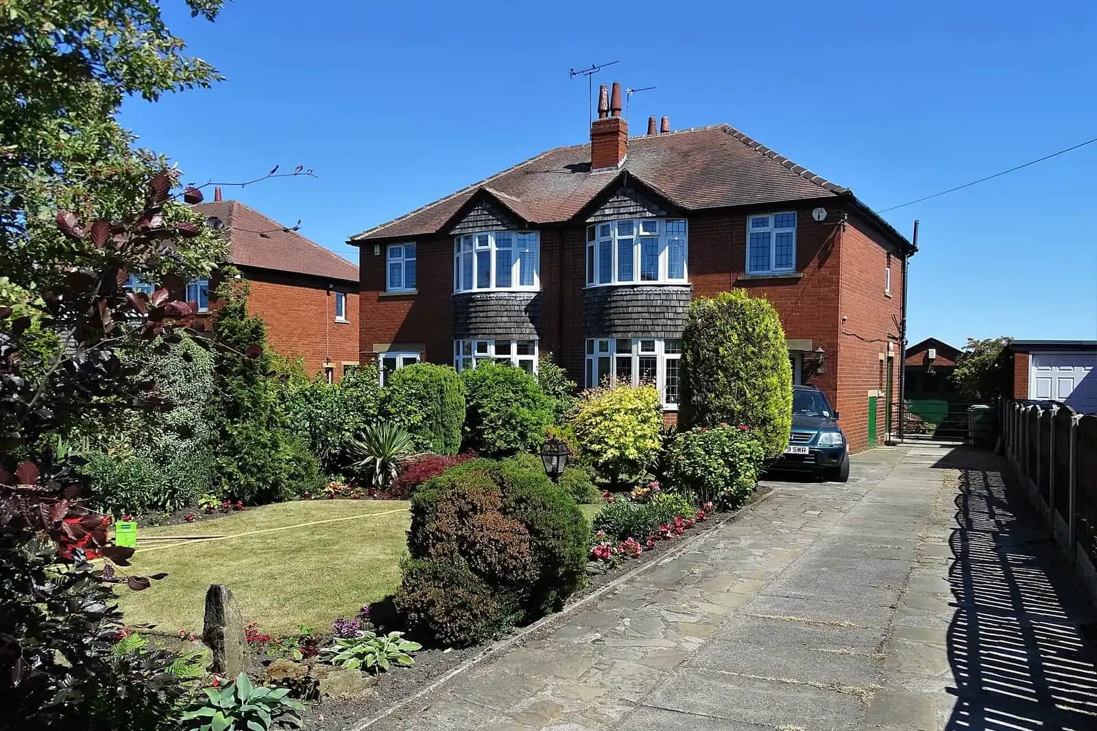 A semi-detached brick house with bay windows and a pitched roof, a stone-flagged drive and a front garden full of shrubs and flower beds on a sunny day