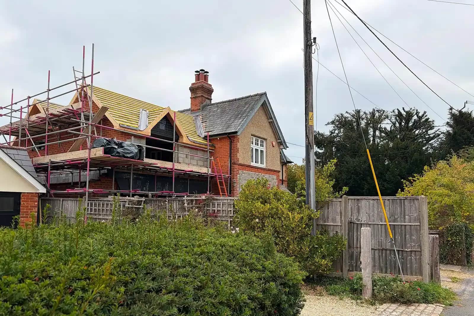 A brick house with a new first-floor roof structure under construction, scaffolding around it and yellow roof membrane on the new frame