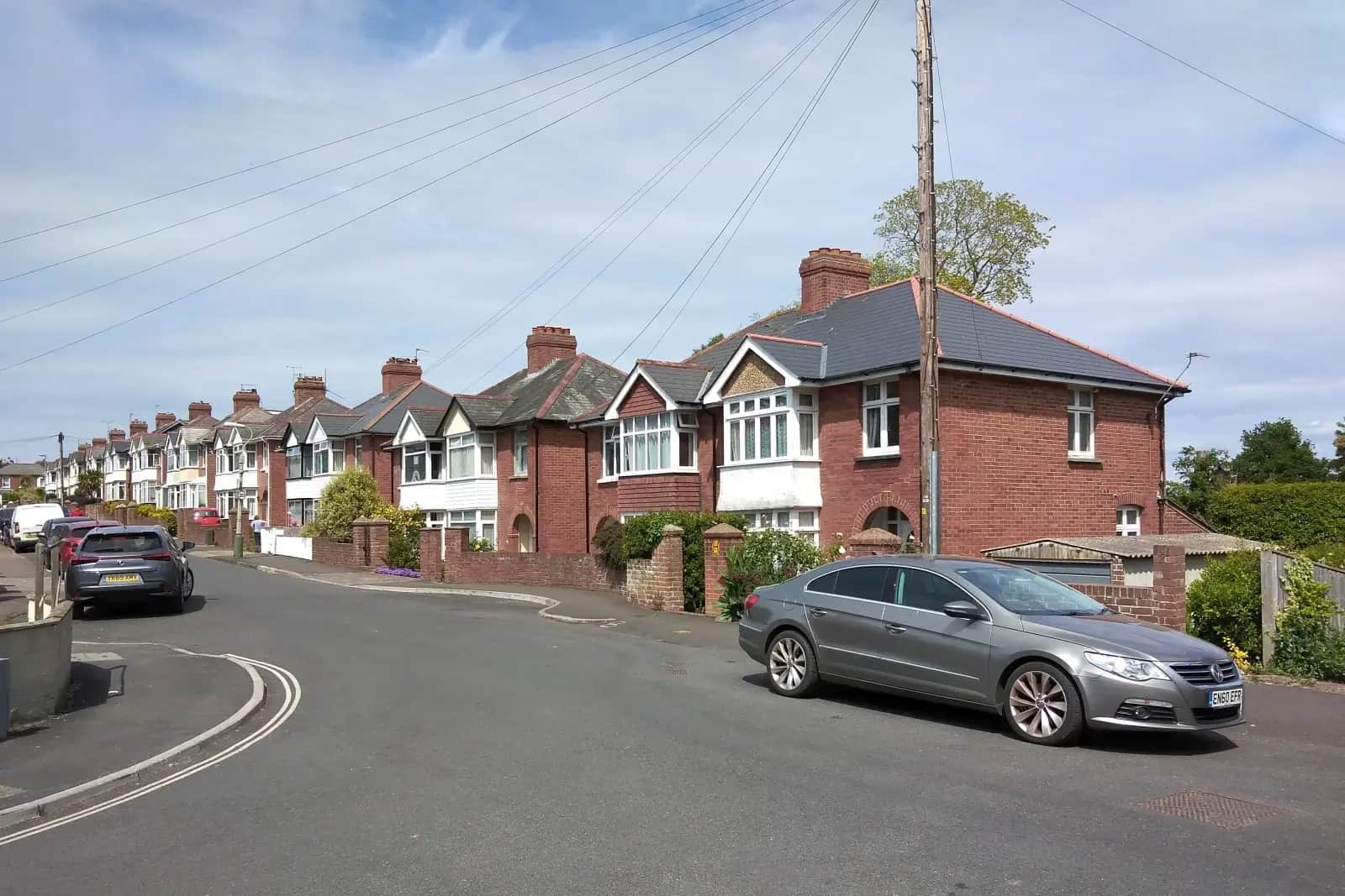 A curving street of 1930s semi-detached houses in red brick and white render in Exeter, a grey car parked in the foreground