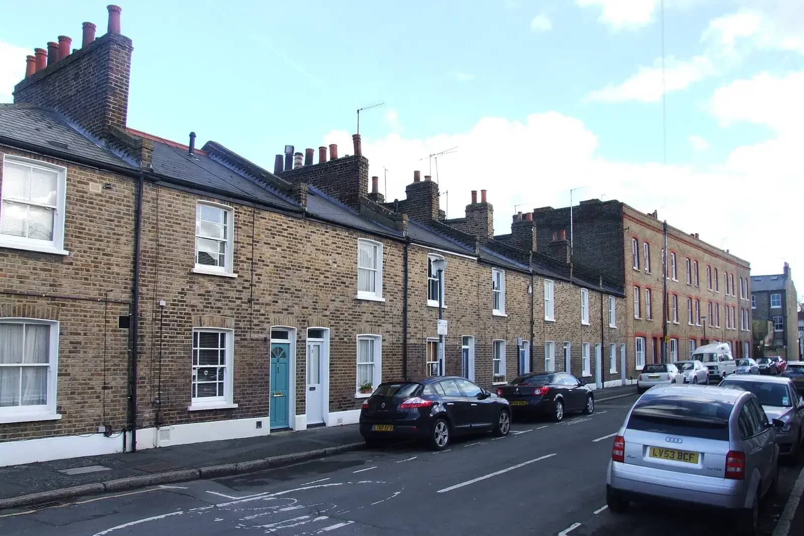 Terrace of small yellow-brick cottages with painted doors on Gibson Street in Greenwich, cars parked along the right