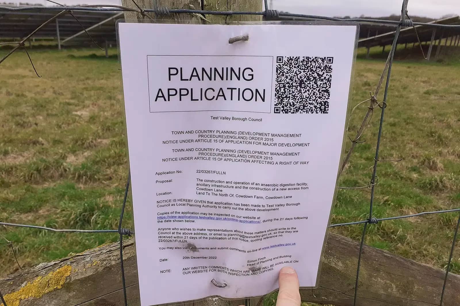 A printed planning application notice with a QR code and the words PLANNING APPLICATION, held by a fingertip against a wire fence in a field