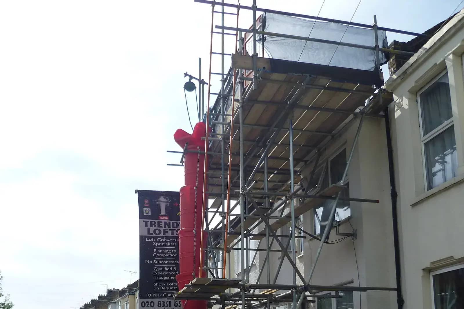 Scaffolding against the front of a white terraced house with a red rubbish chute and a banner for a loft conversion company