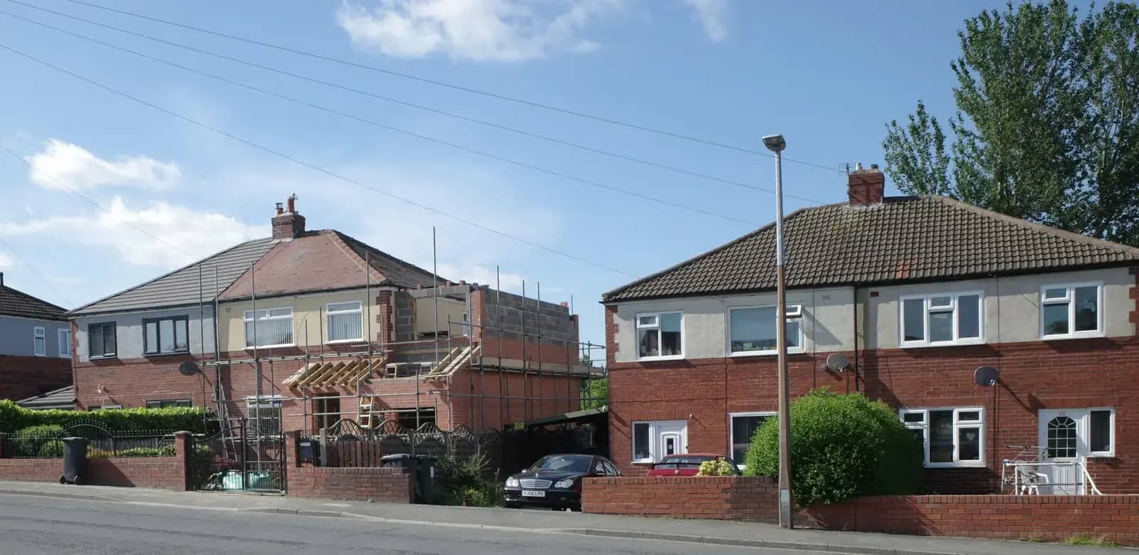 A semi-detached house with a first-floor extension being built, scaffolding and new brickwork showing, beside its unextended neighbour
