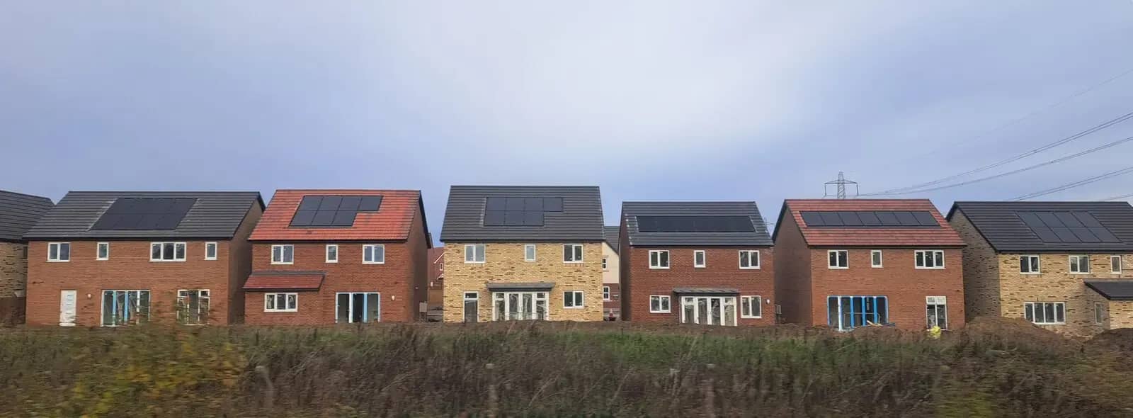 Row of new detached brick houses with solar panels on their roofs, seen across a field from a train