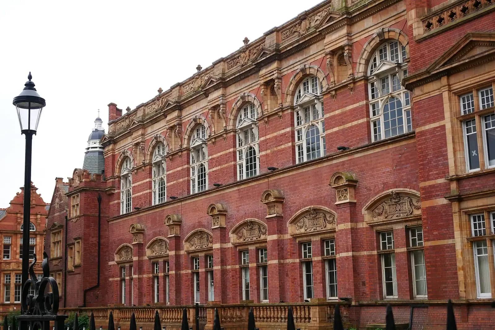 The long red-brick side of a Victorian civic building in East Ham with pale terracotta bands, arched windows and carved pediments, and a black street lamp in front