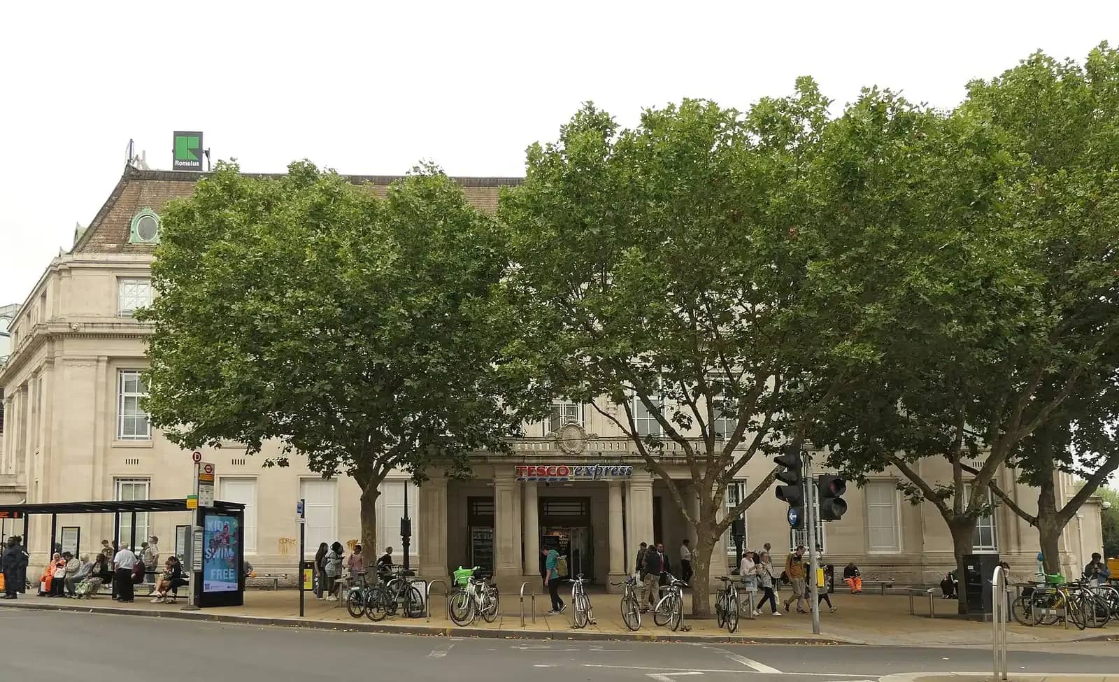 A stone-fronted civic building, Wimbledon Town Hall, behind a row of plane trees with a bus stop and cyclists in front and a supermarket sign over the entrance