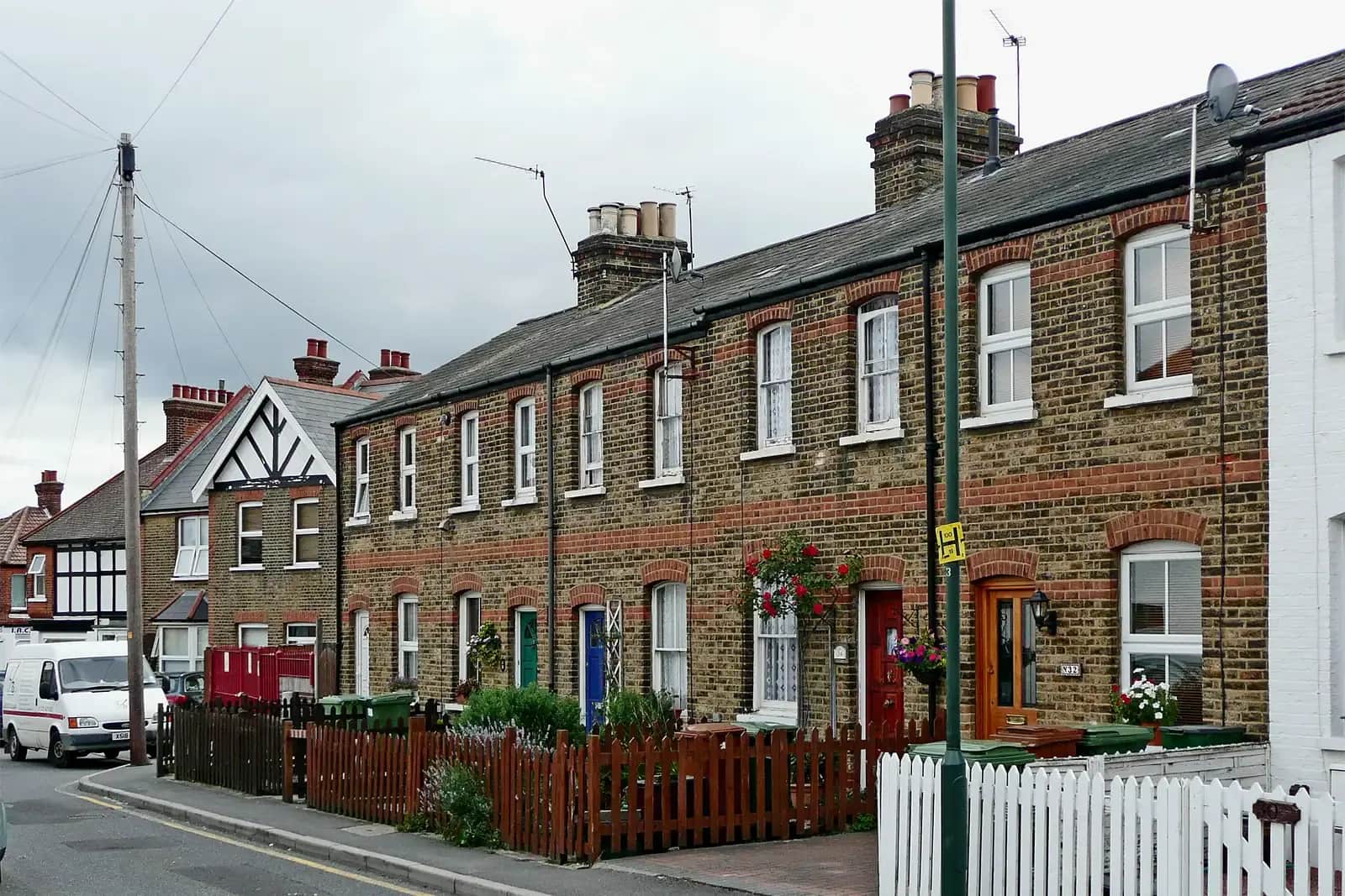 A row of two-storey yellow-brick terraced cottages with red brick banding and white sash windows on a street in Sidcup, front gardens behind picket fences