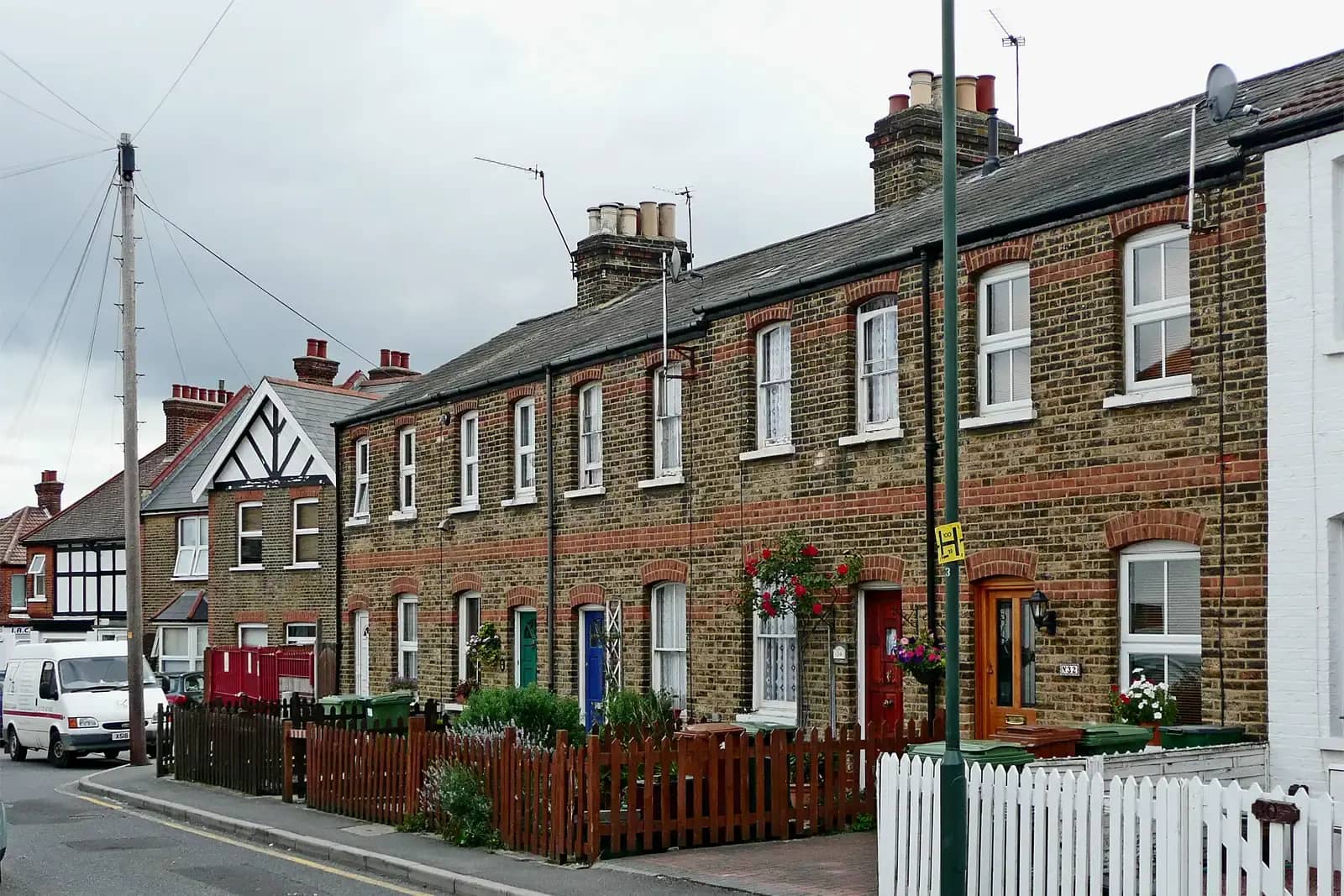 A row of two-storey yellow-brick terraced cottages with red brick banding and white sash windows on a street in Sidcup, front gardens behind picket fences