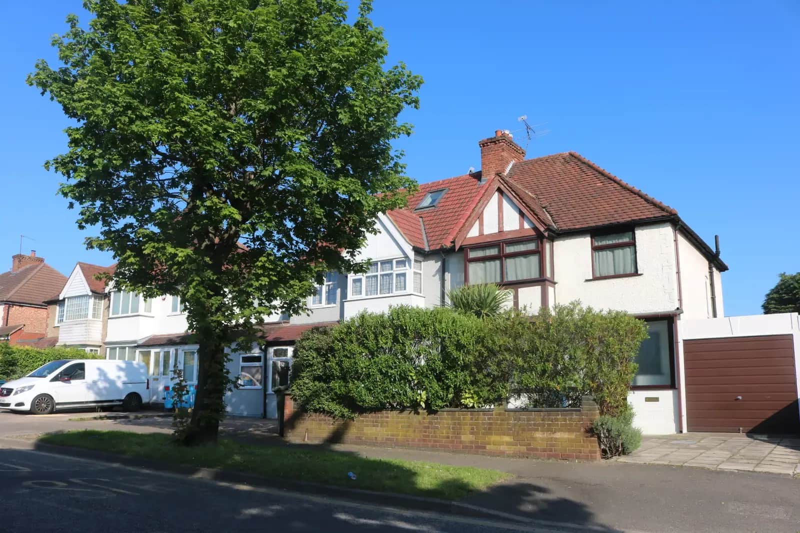 A white-fronted 1930s semi-detached house in North Harrow with a brown up-and-over garage door at its side, a hedge and a large tree in front