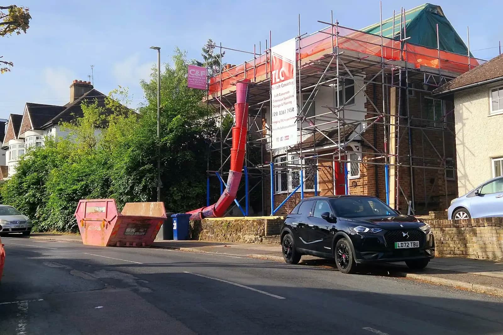 A semi-detached house in East Barnet wrapped in scaffolding and orange sheeting with a red rubbish chute down to a skip, a loft conversion under construction