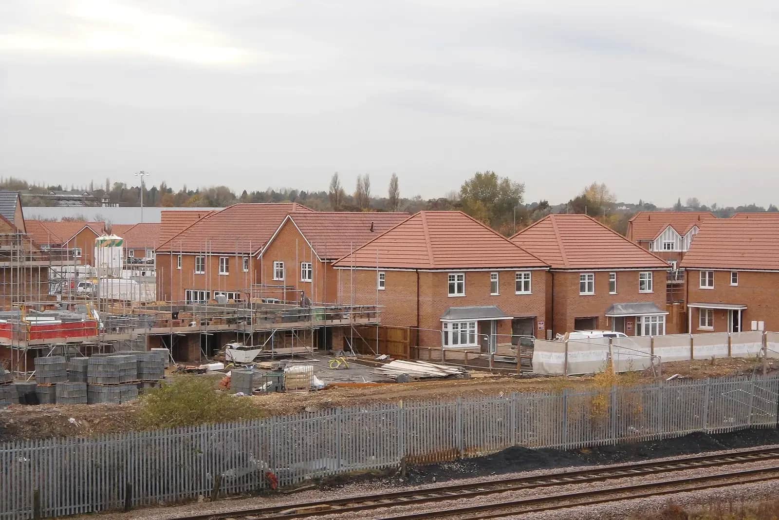 A new-build housing estate of red-brick houses with scaffolding and stacked blocks, seen across a railway line fence