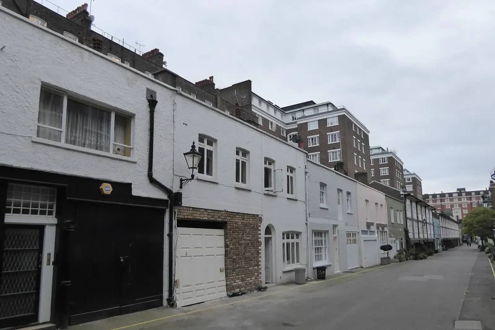London mews of white-painted converted coach houses with black and white garage doors and a gas-style lamp, mansion flats beyond