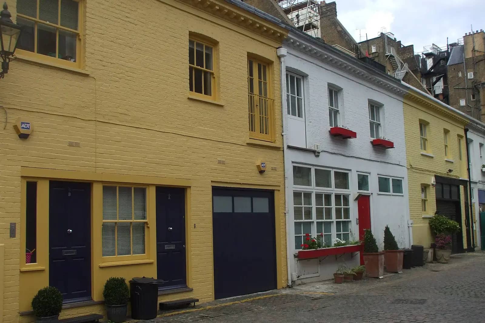 Mews houses painted yellow and white with dark doors, a garage door and red window boxes on a cobbled lane