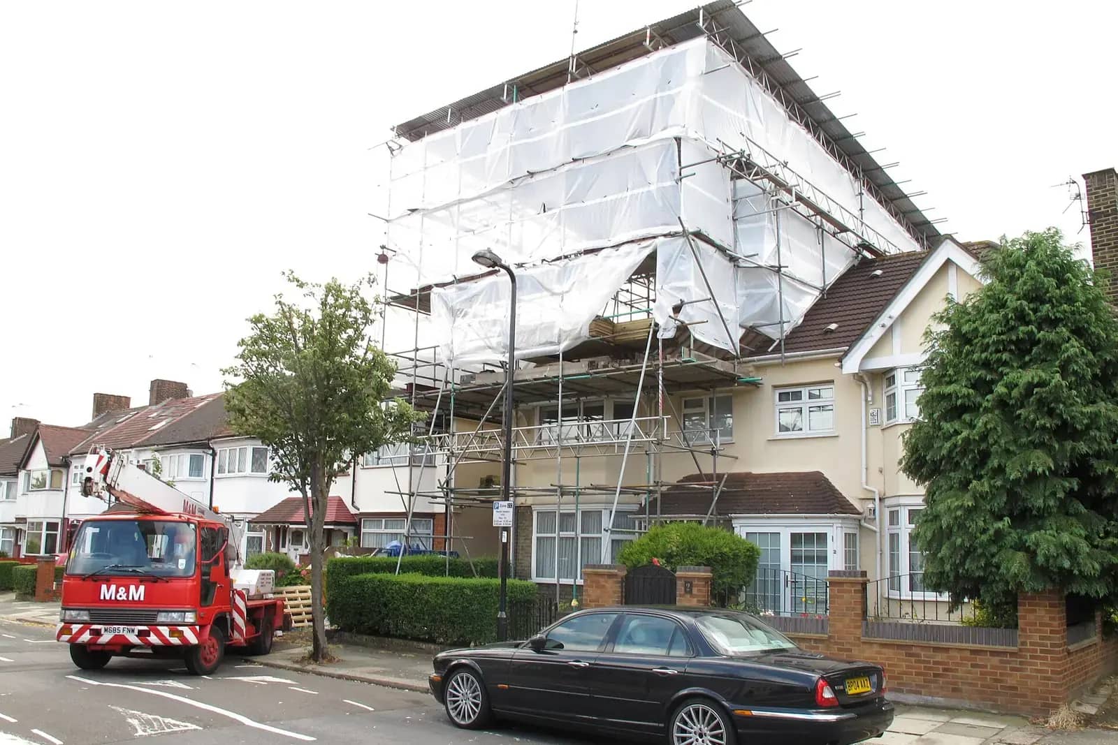 Suburban semi-detached house wrapped in scaffolding and white sheeting for a loft conversion, a red cherry-picker lorry in the road