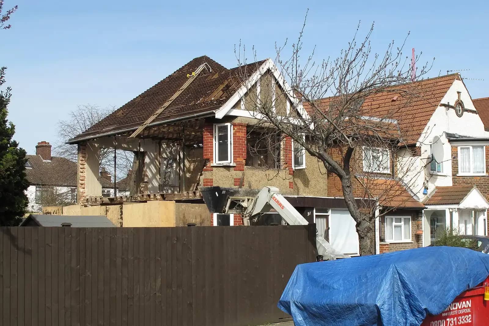 House stripped back for a major extension with its roof timbers exposed, a mini excavator behind the fence and a blue-tarped skip beside it