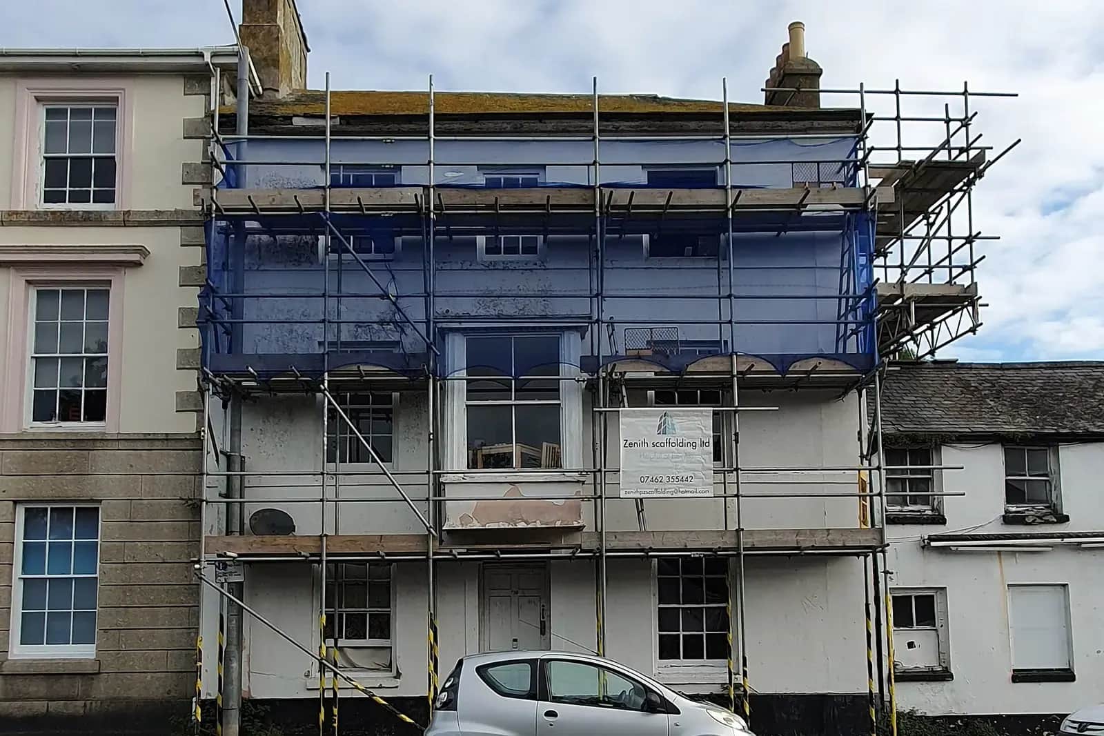 Three-storey white house in a terrace wrapped in blue-netted scaffolding, with an advert board for a scaffolding firm and a small silver car parked below