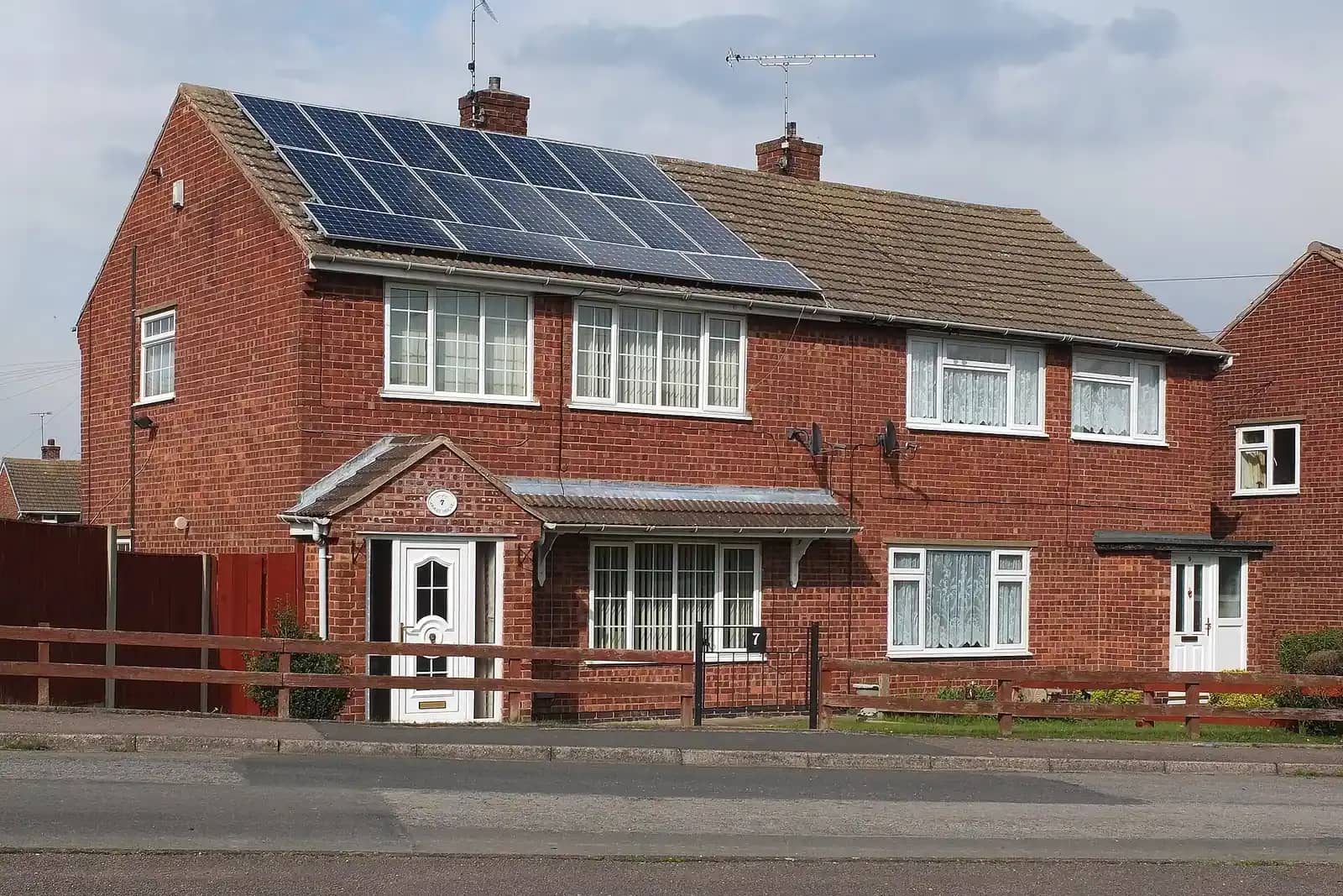 Red-brick semi-detached house with solar panels on its roof, a wooden fence in front and a white front door