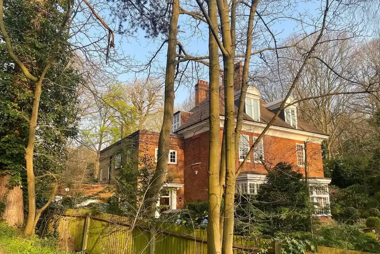 Large red-brick house with dormers seen between tall bare trees on a sunny spring day, a wooden fence in front