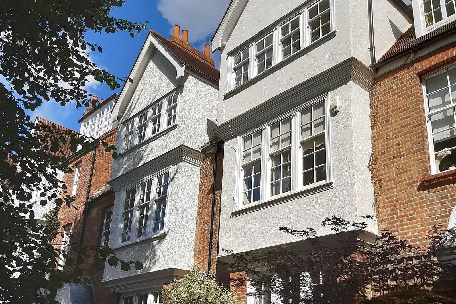 Two tall white roughcast houses with gabled bays and white sash windows between red brick walls, with a Japanese maple in front