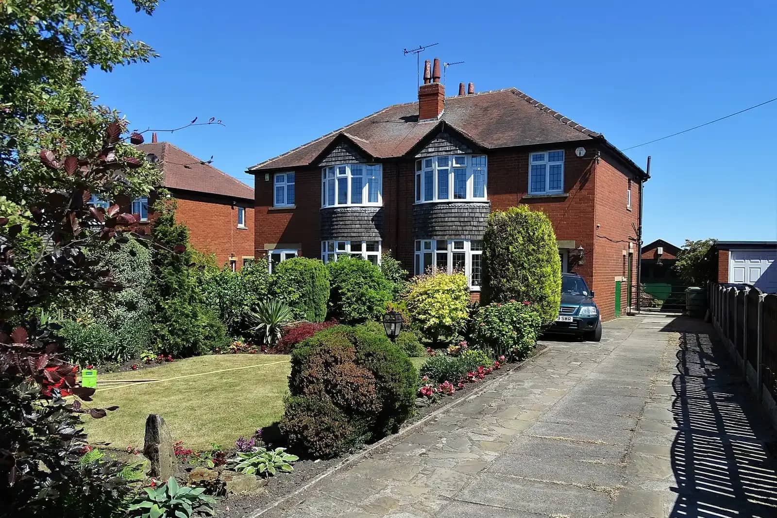 A semi-detached brick house with bay windows and a pitched roof, a stone-flagged drive and a front garden full of shrubs and flower beds on a sunny day