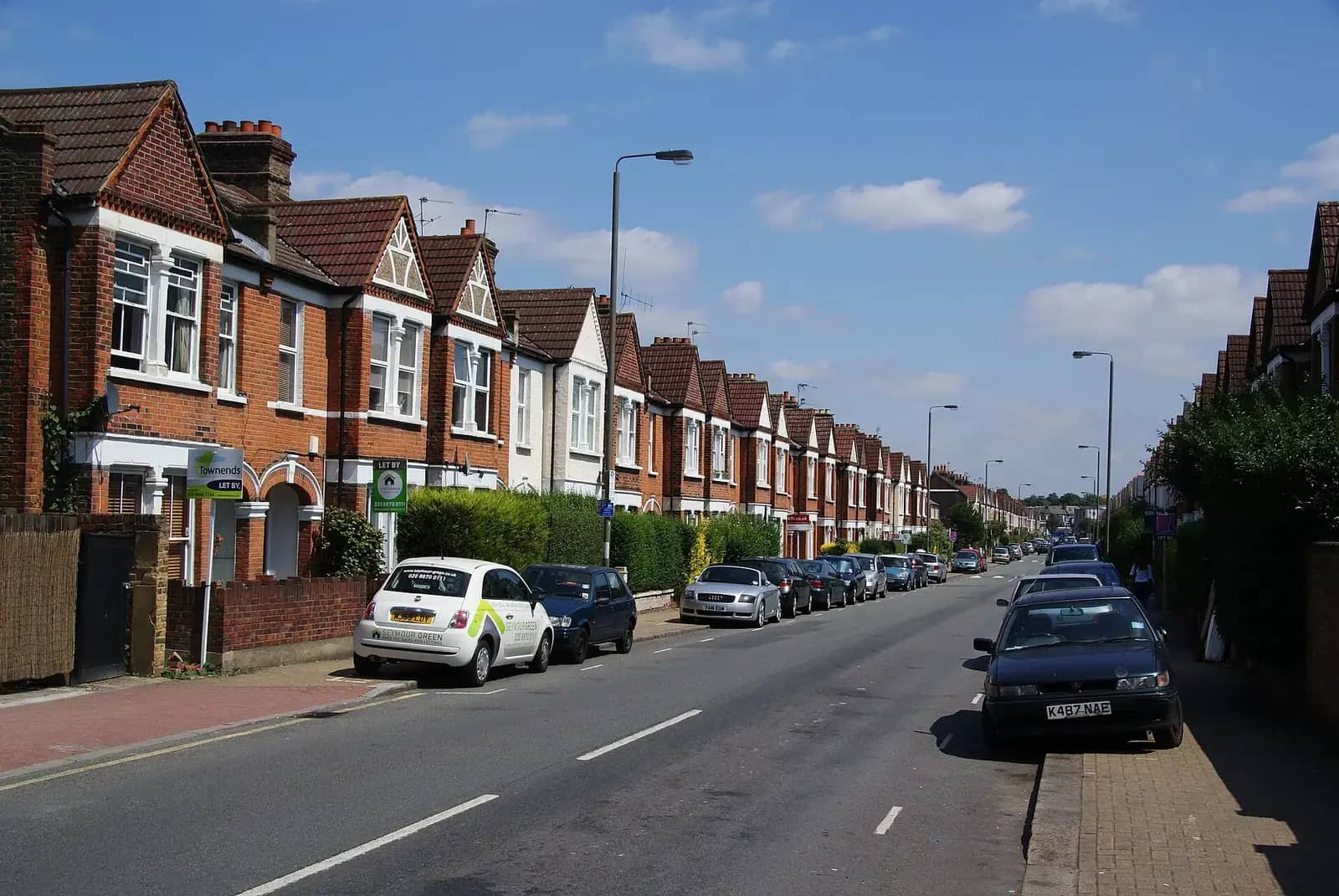 A long street of red-brick Edwardian terraced houses with gables and bay windows, one with a letting board outside, cars parked along the kerb