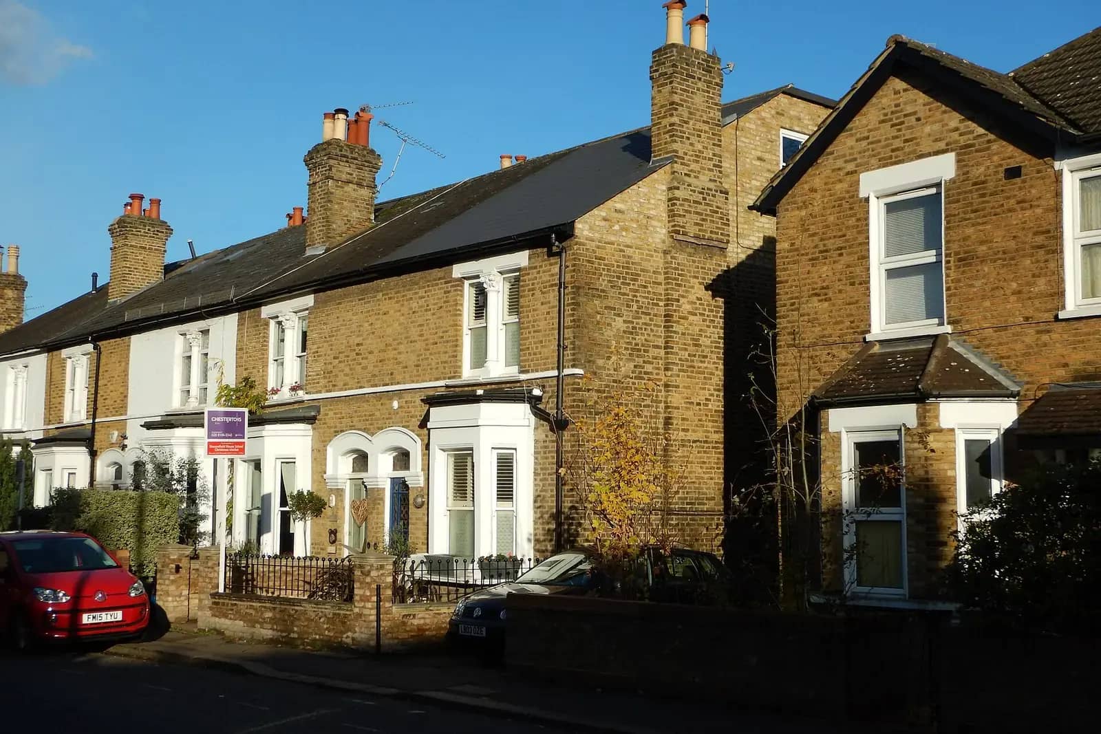 A terrace of yellow-brick Victorian houses with white bay windows, an estate agent board on one front wall and a red car parked at the kerb