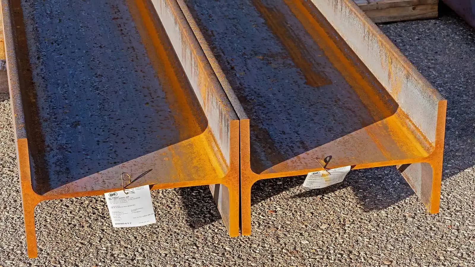 Two rusty steel I-beams lying on gravel with paper labels attached