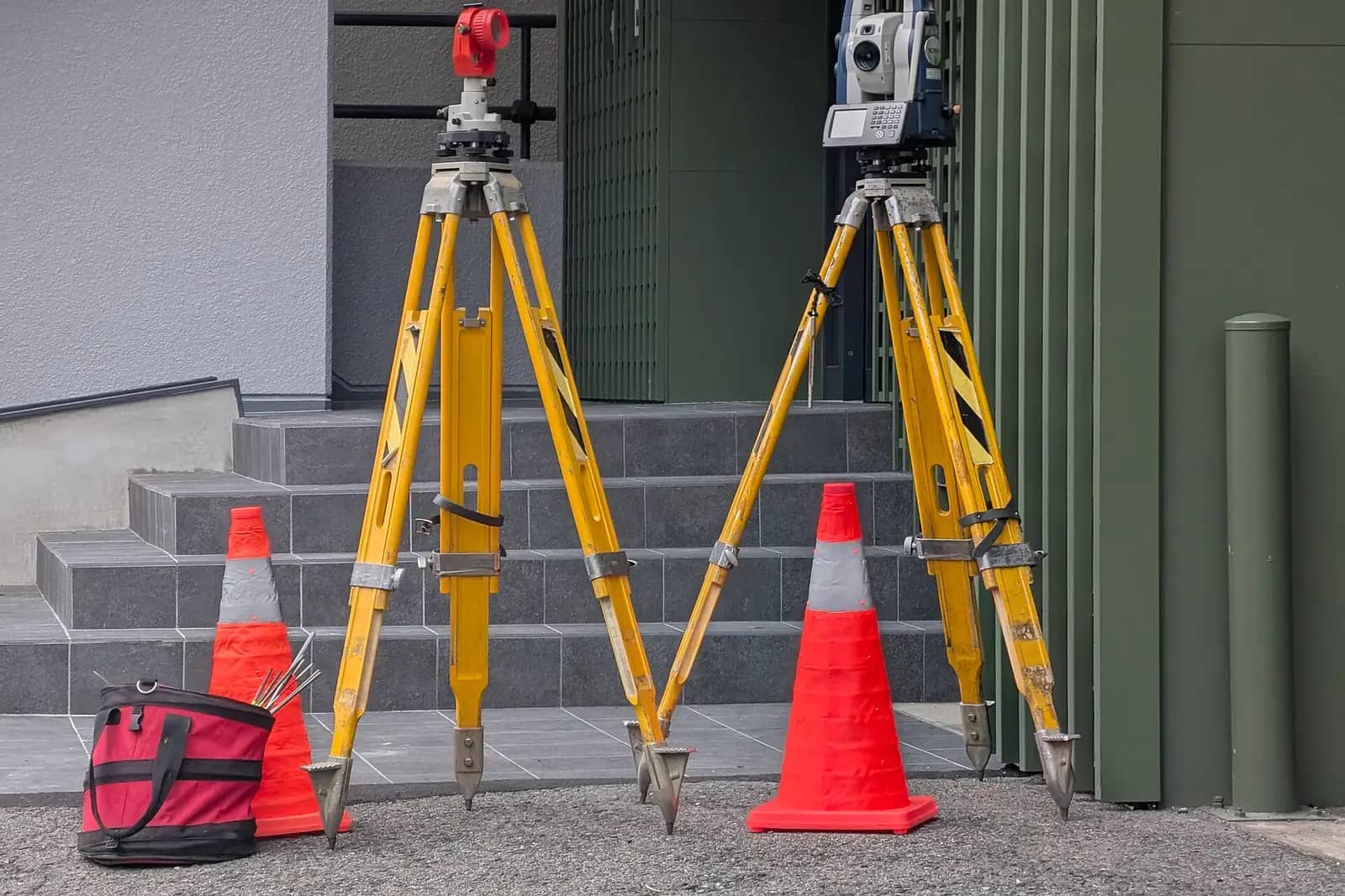 Two yellow survey tripods carrying a total station and a prism, orange cones and a kit bag on a paved path
